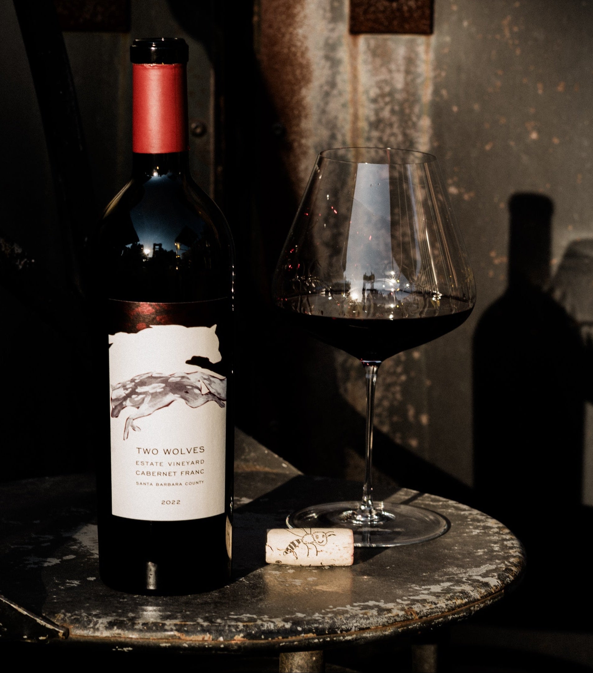 Open bottle of Two Wolves Cabernet Franc  with a wine glass on dark stool.
